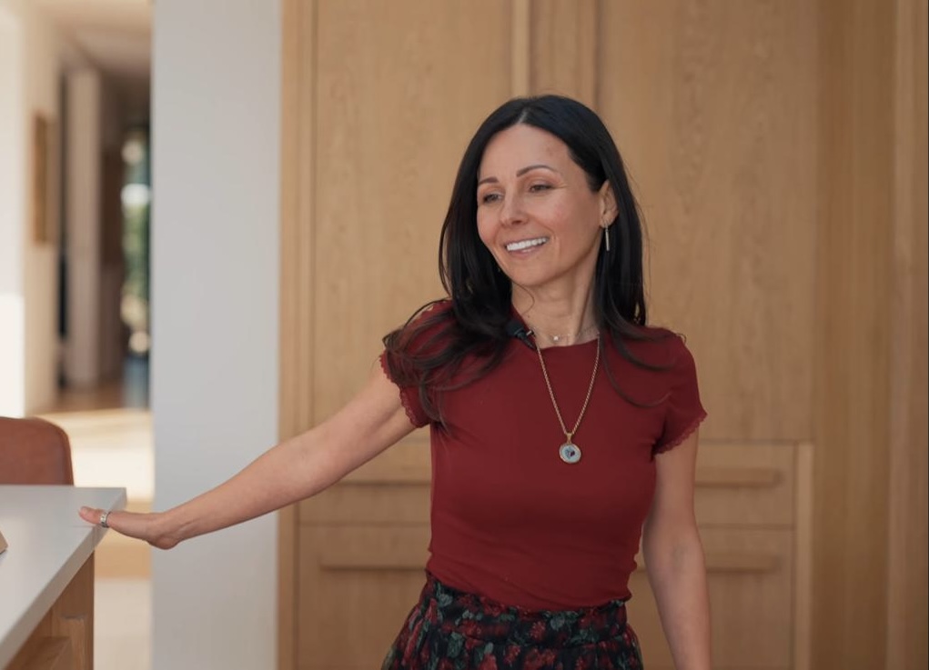Goreti in a red top, her hand resting on a kitchen counter in a bright oak-panelled property.